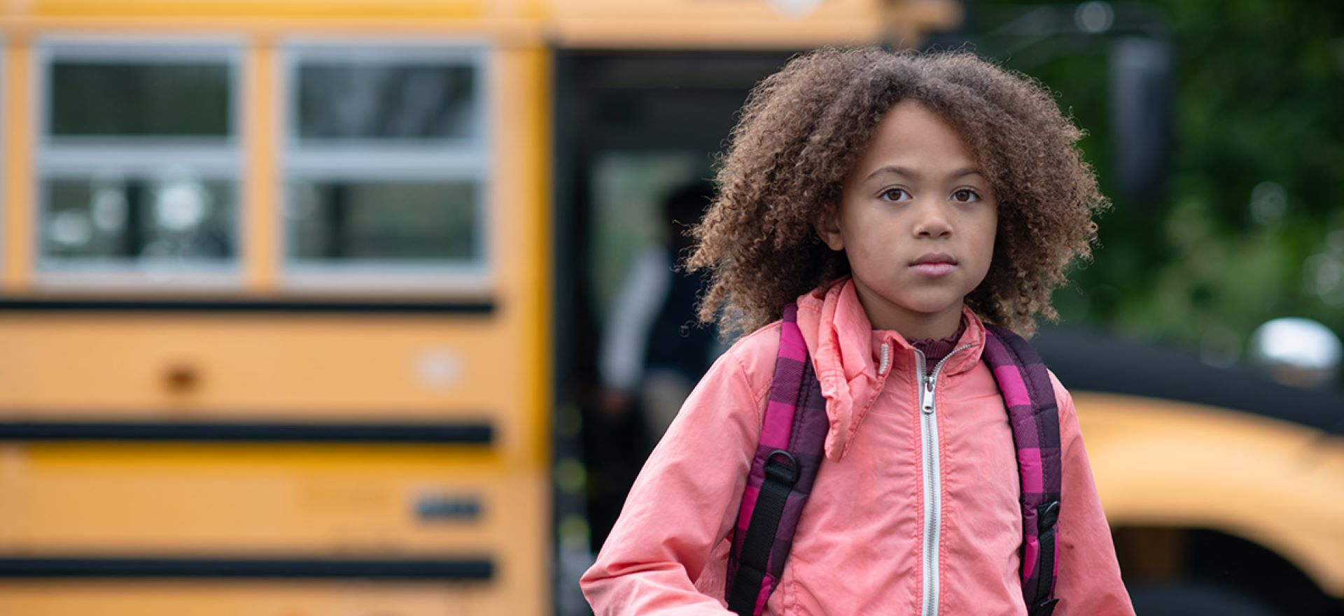 A school-age child, wearing a backpack and a pink jacket, stands in front of a school bus and stares at the camera with a serious expression.